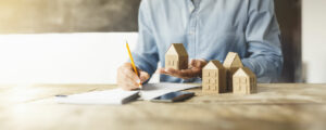 Une personne en chemise bleue tient une petite maquette en carton d'une maison tout en écrivant sur un bloc-notes avec un crayon. Plusieurs modèles de maisons similaires sont disposés sur la table en bois, à côté d'un smartphone.